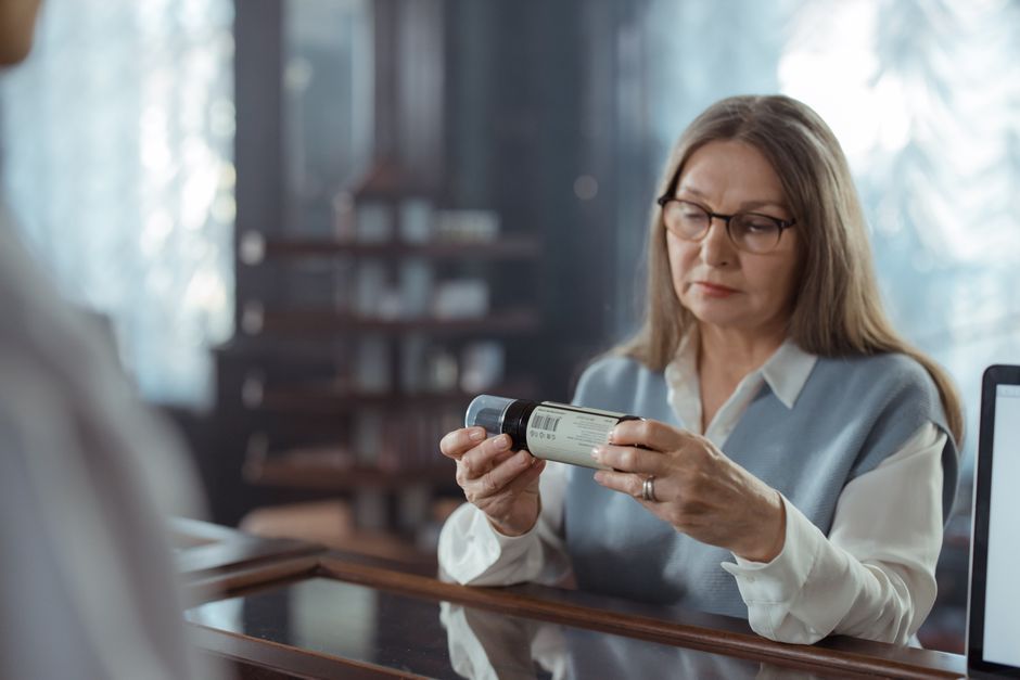A senior woman with eyeglasses reviews a medicine bottle at a pharmacy counter indoors.